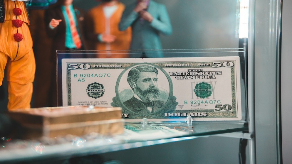 A fifty-dollar bill is displayed in a protective sleeve inside a glass case, conceptually representing the ongoing debate over the Roshtein net worth and the 'Roshtein fake money' allegations, questioning if the money on stream is real or just for show.