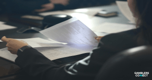 A person in professional attire sits at a conference table reviewing a formal report, representing the UK Gambling Commission's response to the Office for Statistics Regulation's review of the Gambling Survey for Great Britain.