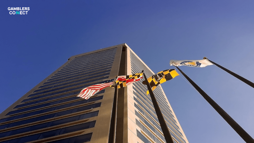 A low-angle photograph capturing a tall, modern office building under a clear blue sky, with the United States flag and the distinctive Maryland state flag prominently displayed on flagpoles. This imagery symbolizes the regulatory authority of the Maryland Lottery and Gaming Control Agency (MLGCA) in approving bet365's sports betting license. The "Gamblers Connect" logo is visible in the top left corner.