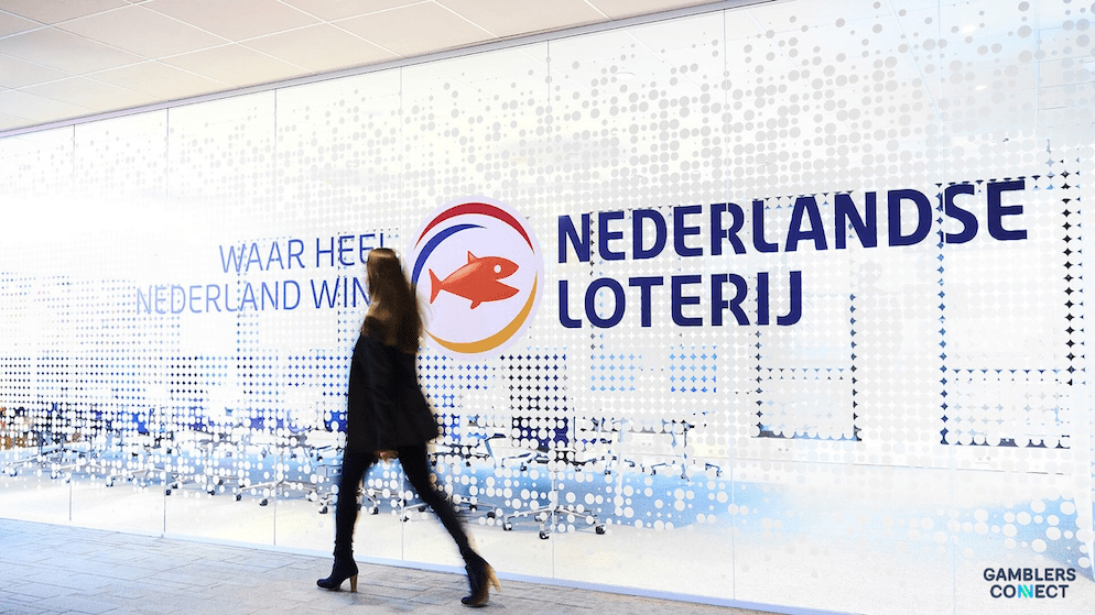 A person in business attire walks inside a modern office, with the prominent "Nederlandse Loterij" logo and "WAAR HEEL NEDERLAND WINT" slogan displayed on a glass wall.