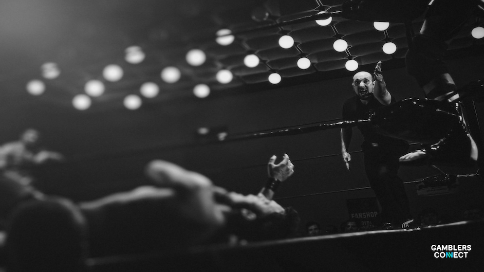 A black and white photo capturing the chaotic scene in a wrestling ring. A wrestler is down on the mat as another person stands over him, with others rushing in from the side.