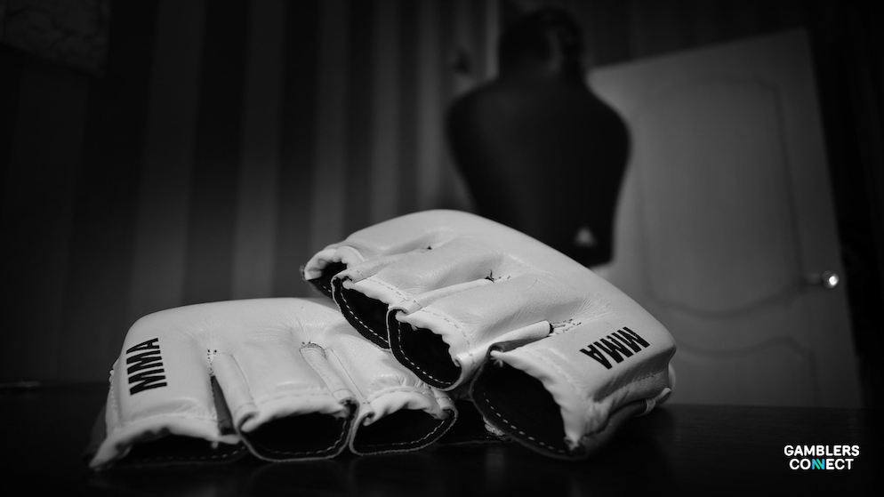 A black and white close-up of a pair of white open-fingered MMA gloves with black accents lying on a reflective dark surface. The word "MMA" is clearly visible in black lettering on the wrist strap of the glove in the foreground. The background is softly blurred, showing a figure standing near a doorway, subtly hinting at the world of mixed martial arts.dd