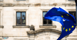 A vibrant blue European Union flag with yellow stars, billowing in front of a classical European building, symbolizing the EGBA's call for a new, harmonized standard on markers of harm in online gambling across Europe.