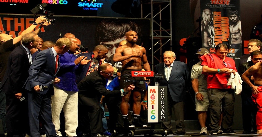 Floyd Mayweather Jr. on the scale during a weigh-in for a major boxing event, surrounded by officials and media, symbolizing the preparation for the record-breaking pay-per-view fights that defined his career and built his substantial Floyd Mayweather net worth.