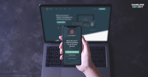 A hand holding a smartphone displaying the Gamban app, which blocks online gambling, with a laptop in the background showing the Gamban website, symbolizing the software's multi-device protection and new availability in Japanese.