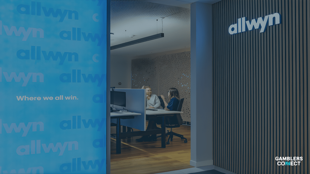 Two Allwyn employees discussing work at a desk in a contemporary office setting, representing the team behind the UK National Lottery's technology and terminal upgrades.