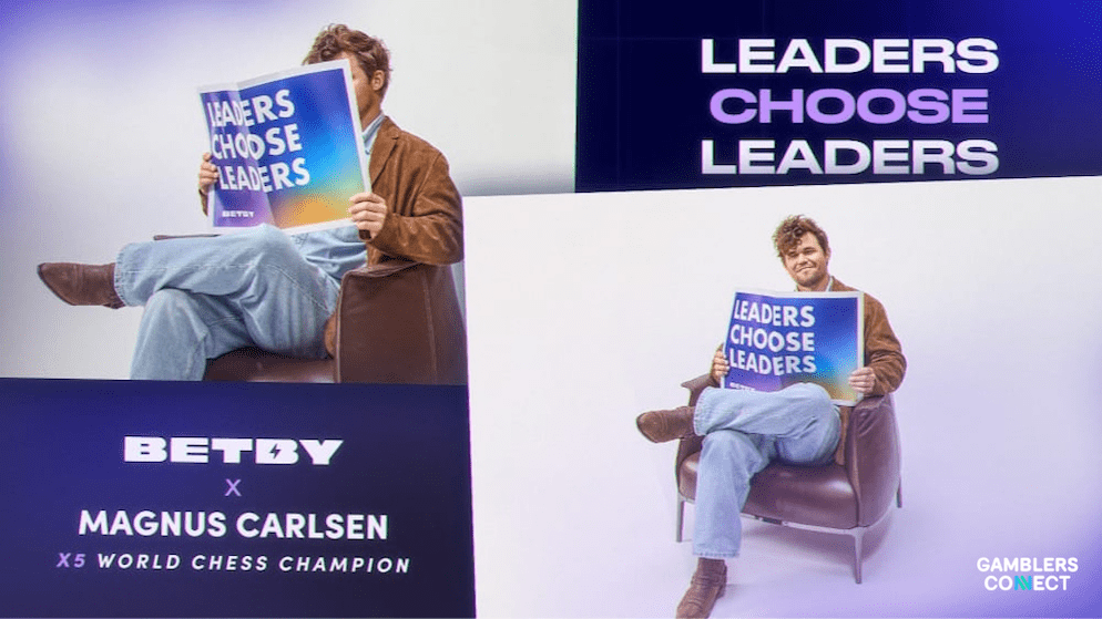 Magnus Carlsen, 5-time World Chess Champion, seated and holding a sign that reads "Leaders Choose Leaders" for the Betby campaign, with campaign branding visible.