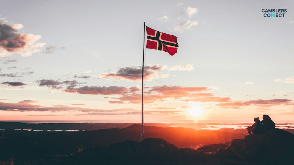 The Norwegian flag waving against a dramatic sunset, with people silhouetted in the foreground, representing the Norwegian Lottery Authority's fine against Norsk Tipping for a Eurojackpot prize error.