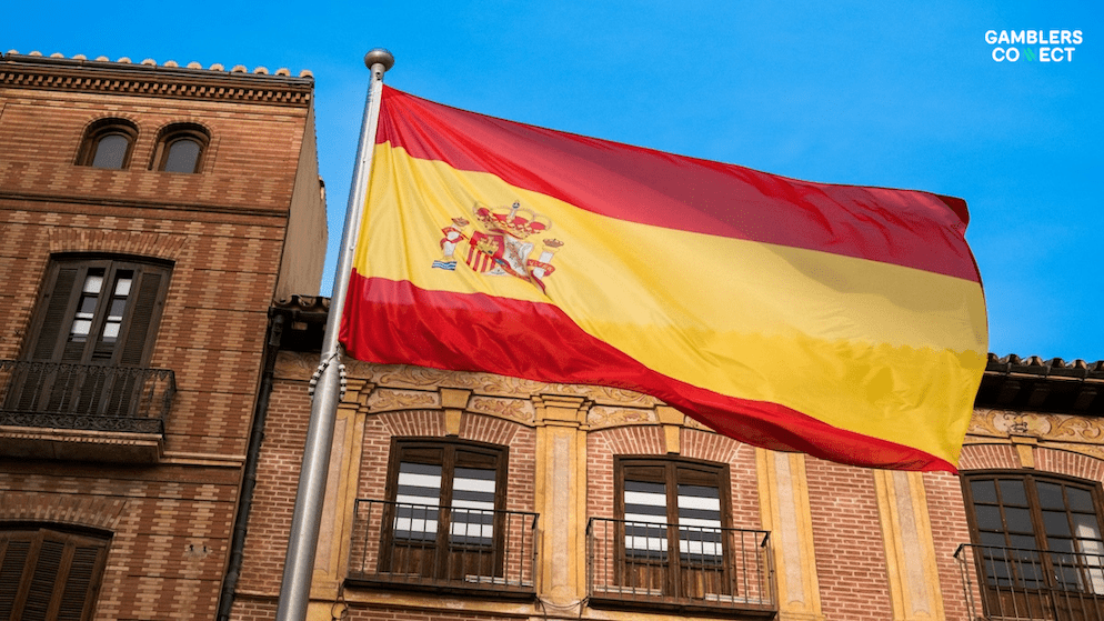 The flag of Spain, with its coat of arms, flying against a blue sky next to traditional Spanish architecture, representing the nation's new public awareness campaign by the Ministry of Consumer Affairs.