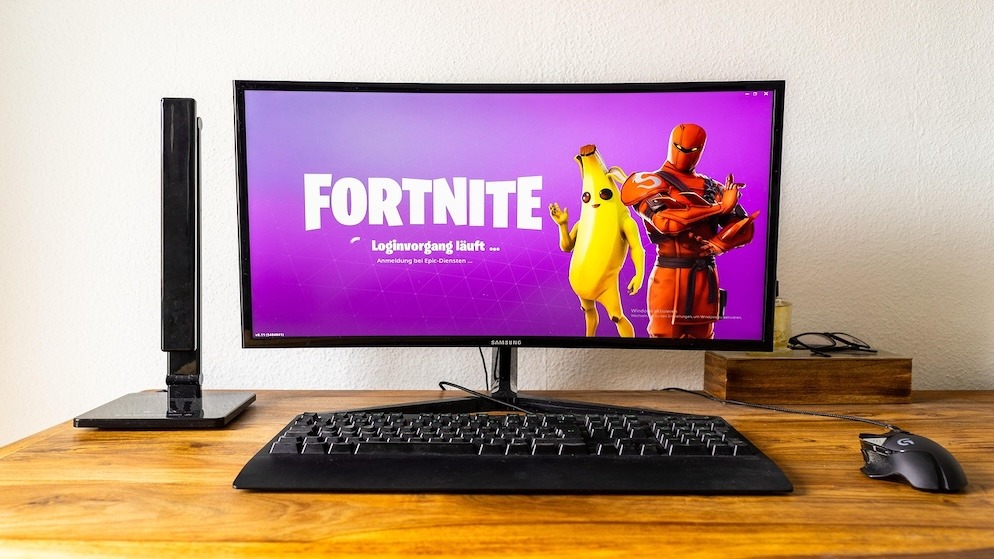 A desktop monitor displaying the Fortnite login screen with the characters Peely and Red Knight, symbolizing the starting point of the game's cosmetic economy.