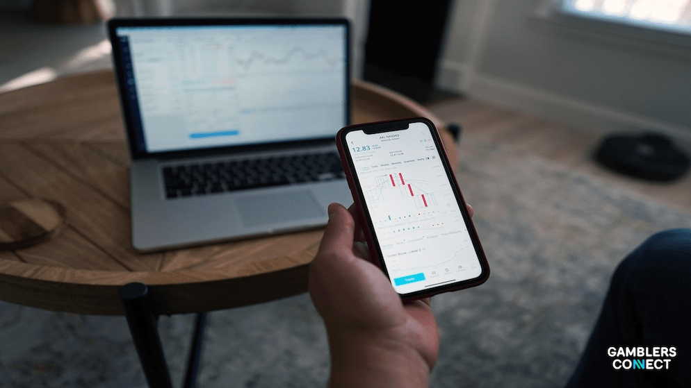 A hand holding a smartphone displaying a financial chart, with a laptop also showing data in the background, symbolizing financial tracking in sports betting - Illinois Gaming Board.