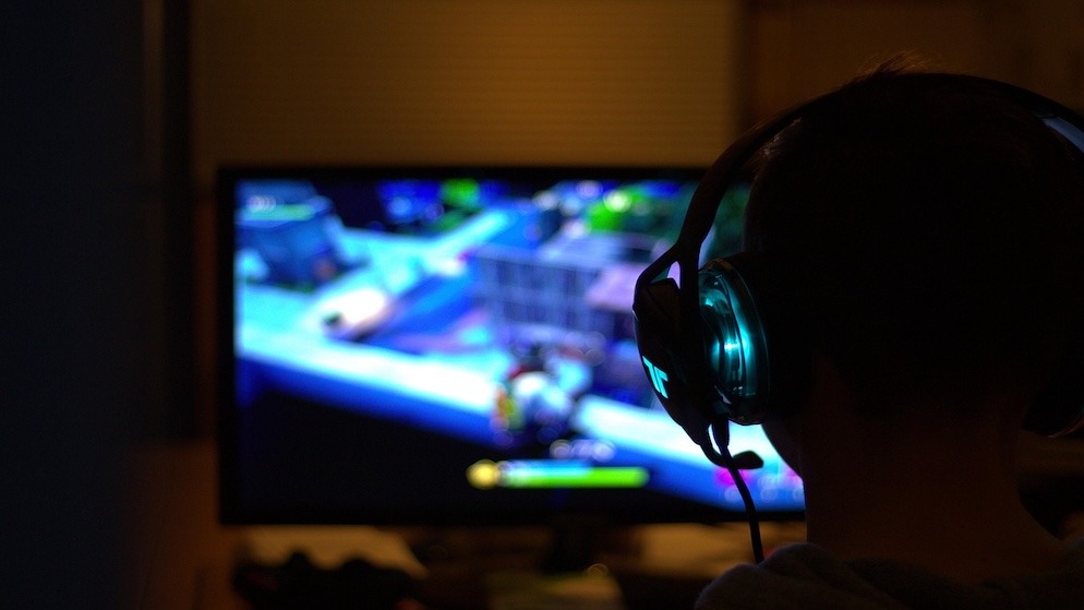 Rear view of a young person wearing illuminated headphones, focused intently on a blurred Fortnite game screen in a dark room, highlighting the target demographic for gambling scams.
