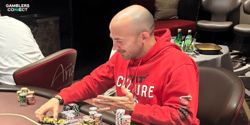 Professional poker player Jason Mercier in a red hoodie, actively playing poker at a live casino table, handling chips.