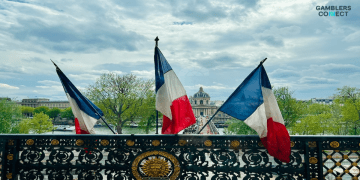 Three French flags waving on a balcony overlooking a city street and a classic building, symbolizing the authority of the French regulatory body, the ANJ.
