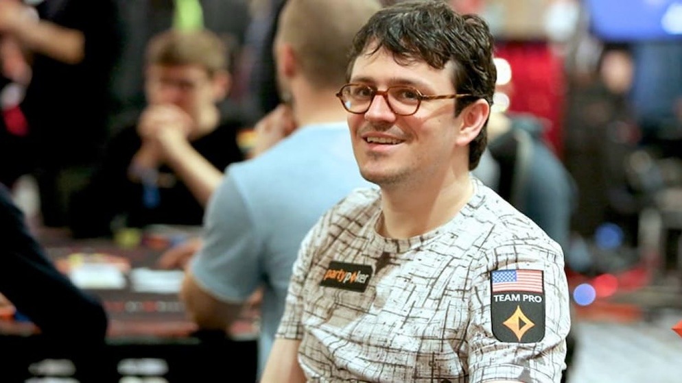 A smiling Isaac Haxton wearing his signature round glasses and a white patterned t-shirt with a "partypoker TEAM PRO" patch on the sleeve, seated at a poker table.
