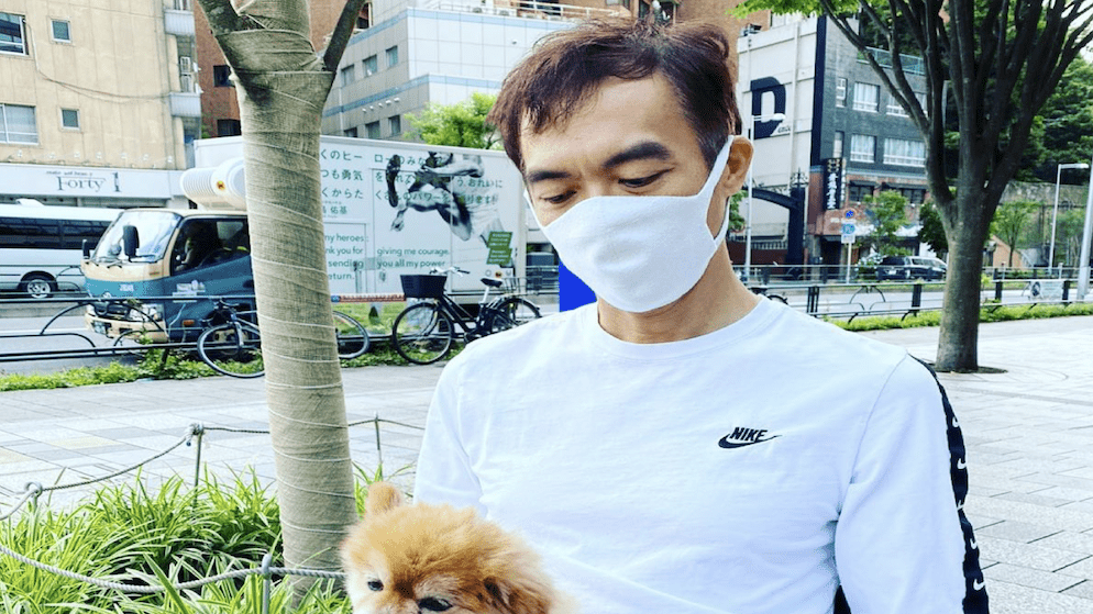 John Juanda wearing a white mask and white shirt, holding a small dog on a street in Tokyo, Japan.