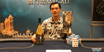 ohn Juanda smiling at a poker table with a large gold trophy and a bottle of Ace of Spades champagne after winning the Triton Macau Main Event.