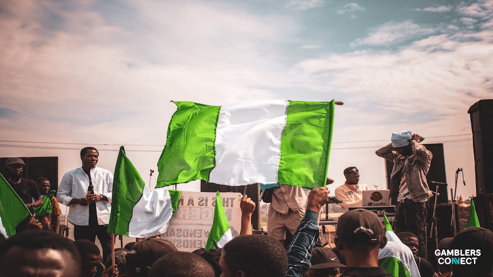 An editorial graphic showing a smartphone displaying a sports betting app interface, superimposed over a blurred image of the Nigerian National Assembly building Central Gaming Bill.