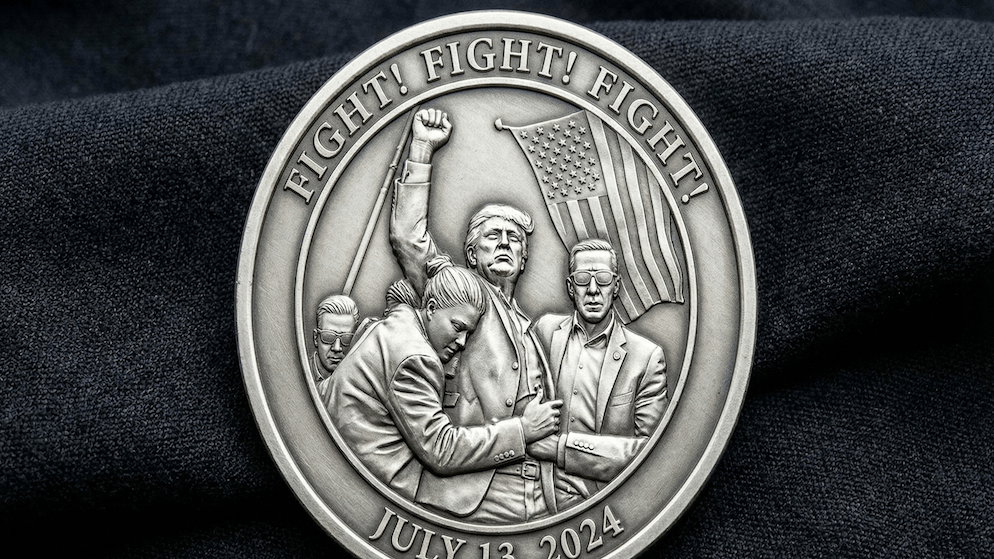 A close-up of a silver commemorative coin featuring Donald Trump with his fist raised and an American flag, surrounded by Secret Service agents, commemorating the Butler, PA rally.