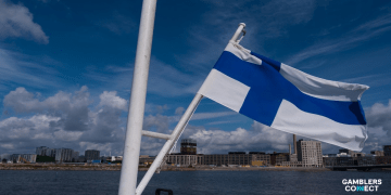 The national flag of Finland flying on a white pole against a blue sky and cityscape, marking the country's transition from a monopoly to a licensed gambling market.