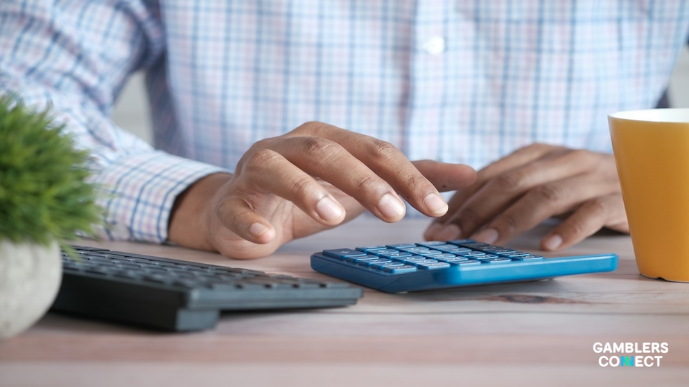 A person using a blue calculator on a wooden desk next to a keyboard and a cup of coffee, representing the calculation of Brazil's BRL 1.03 billion gaming tax from the online gambling market.