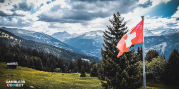 A wide-angle view of a valley in the Swiss Alps featuring a traditional wooden cabin and a Swiss flag waving in the foreground.