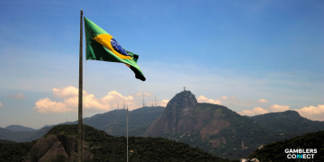 The national flag of Brazil waving on a pole with the iconic Christ the Redeemer statue visible on a distant mountain peak.
