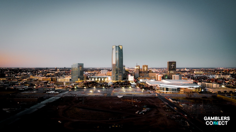 An aerial dusk view of the Oklahoma City skyline featuring the Devon Tower and surrounding downtown buildings under a darkening sky.