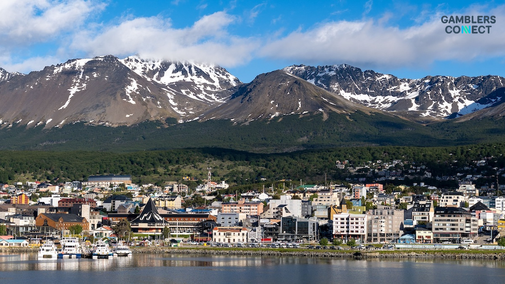 A scenic wide shot of the coastal city of Ushuaia, Argentina, with residential buildings along the water in front of snow-capped mountains under cloudy skies.