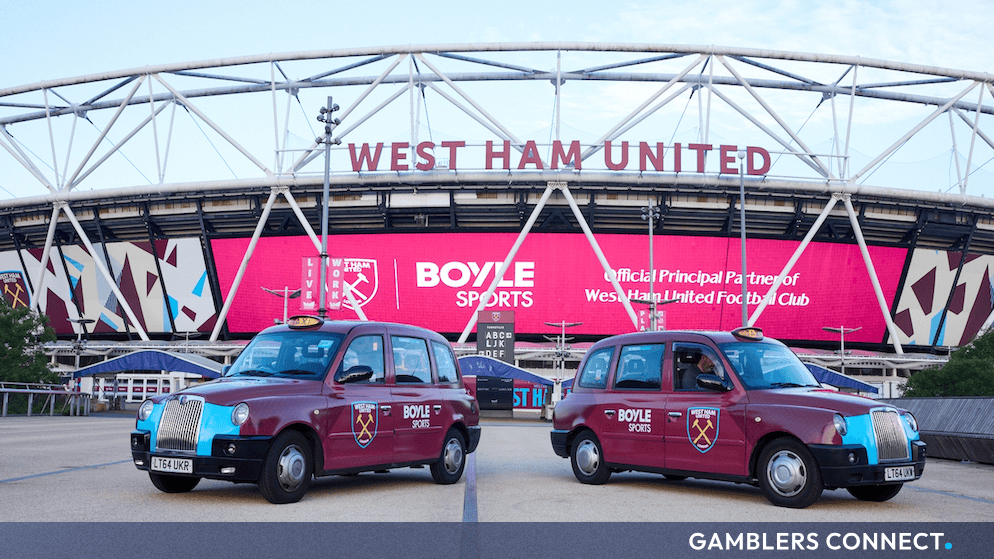 Branded claret and blue promotional taxis parked outside the West Ham United home stadium underneath a large digital sponsorship display.