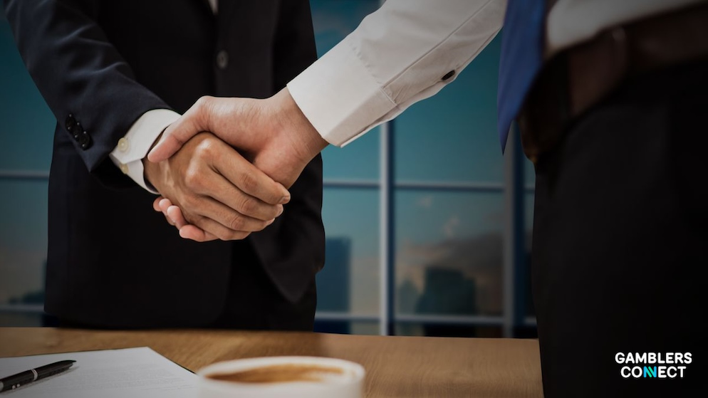 Executive business partners in professional suits shaking hands over a conference table with a city window background view.