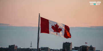 The national flag of Canada—displaying its red vertical bars flanking a central white square containing a single stylized red maple leaf—waving from a pole over a blurred urban cityscape at sunset.