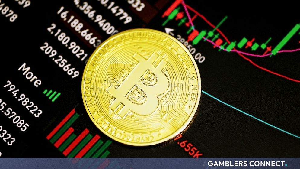A physical golden Bitcoin token positioned on a dynamic stock market trading monitor showing shifting financial indicators.