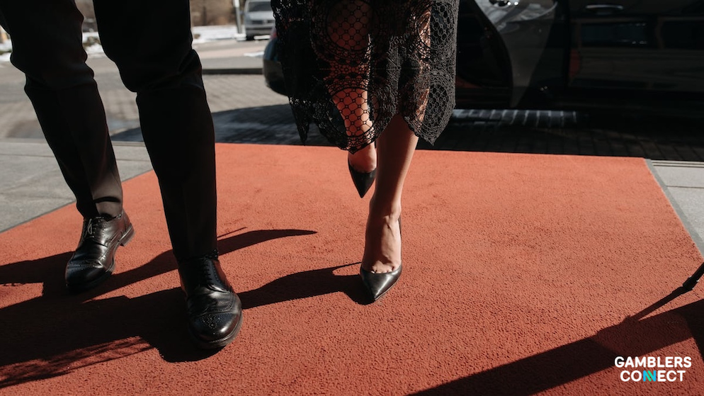 A close-up cinematic focus shot from the ground level showing a man in black dress shoes and a woman in elegant black high heels walking side-by-side along a red carpet walkway next to a vehicle.