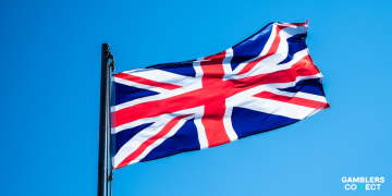 The national flag of the United Kingdom—the iconic red, white, and blue Union Jack—waving prominently from a black metal pole against a solid, cloudless blue sky backdrop canvas.