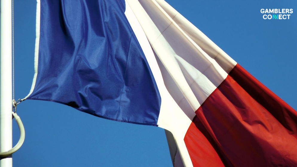 The French national flag, displaying its bold blue, white, and red vertical panels, billowing on a white flagpole against a clear sky.