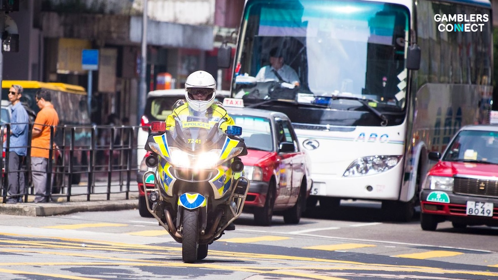 A close-up, ground-level tracking shot of a Hong Kong police officer riding a modern white and blue municipal law enforcement motorcycle down a busy urban road, with a large white double-decker bus and red taxicabs visible in the background.