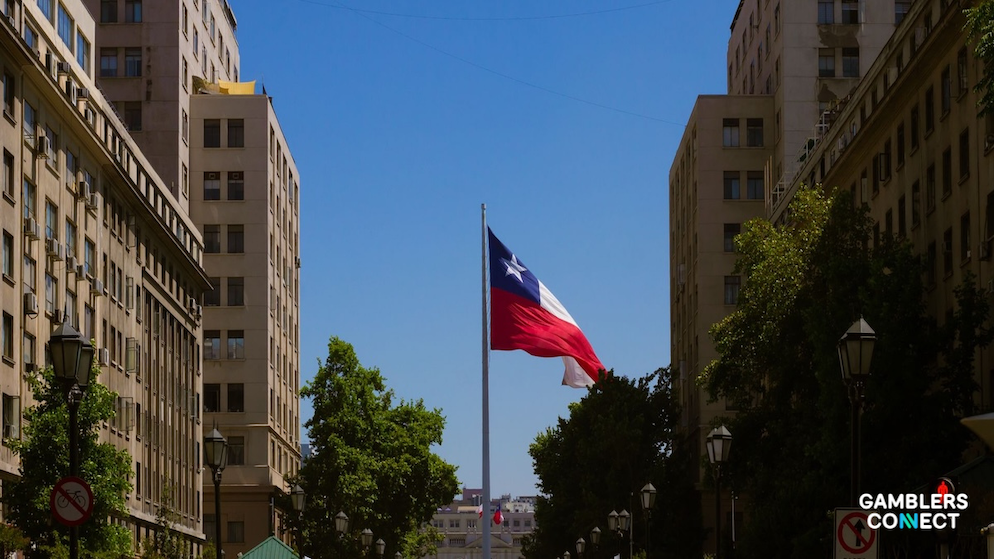 The national flag of Chile flying high on a pole positioned between classical urban apartment buildings in downtown Santiago under a bright blue sky.