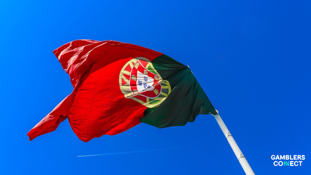 A close-up focus shot of the red and green flag of Portugal rippling under bright sunlight, with a faint white vapor trail visible in the sky.