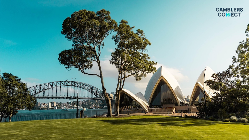 The iconic architectural white shells of the Sydney Opera House viewed from a public park with mature trees under a bright blue sky.