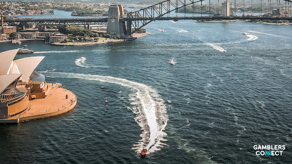 An aerial high-angle shot overlooking the deep blue waters of Sydney Harbour, showing multiple passenger ferries making white water wakes near the Sydney Opera House under a steel bridge.