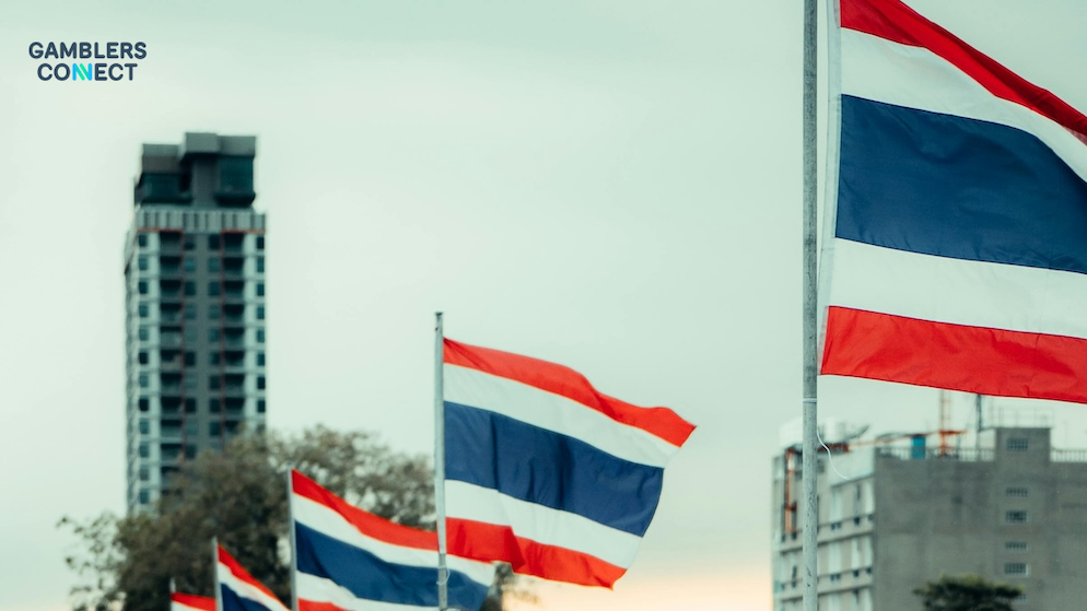 The official Thai national flags waving in the wind with urban high-rise concrete properties visible in the distance under an overcast sky.