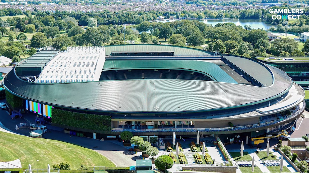 An aerial panoramic view of the iconic green-roofed Centre Court stadium building at Wimbledon, surrounded by manicured lawns, walkways, and tennis facilities under a clear sky.