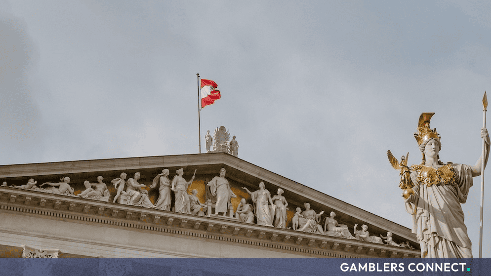 The architectural detail of the Austrian Parliament building roof line, featuring classical stone carvings and the national flag flying under an overcast sky.