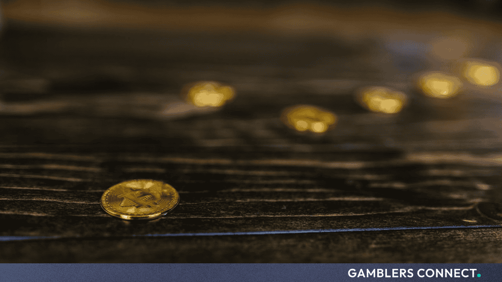 A close-up shot of a physical gold Bitcoin token resting flat on a dark, rustic wood-grained surface, with several other blurred gold tokens extending into the shallow-focus background.