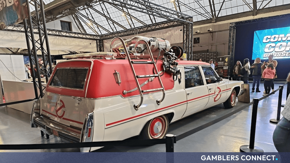 Side profile view of the white and red Ghostbusters vehicle on a convention floor, highlighted under bright overhead exhibition hall lights.