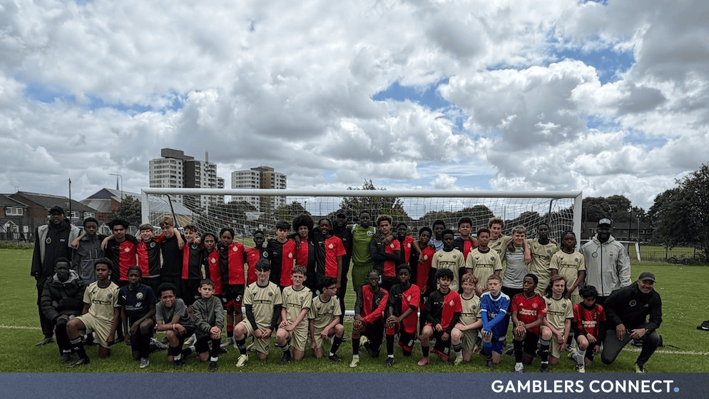Group of young football players and coaches posing on a grass pitch in front of a goalpost, representing grassroots sports supported by MERKUR Casino UK.