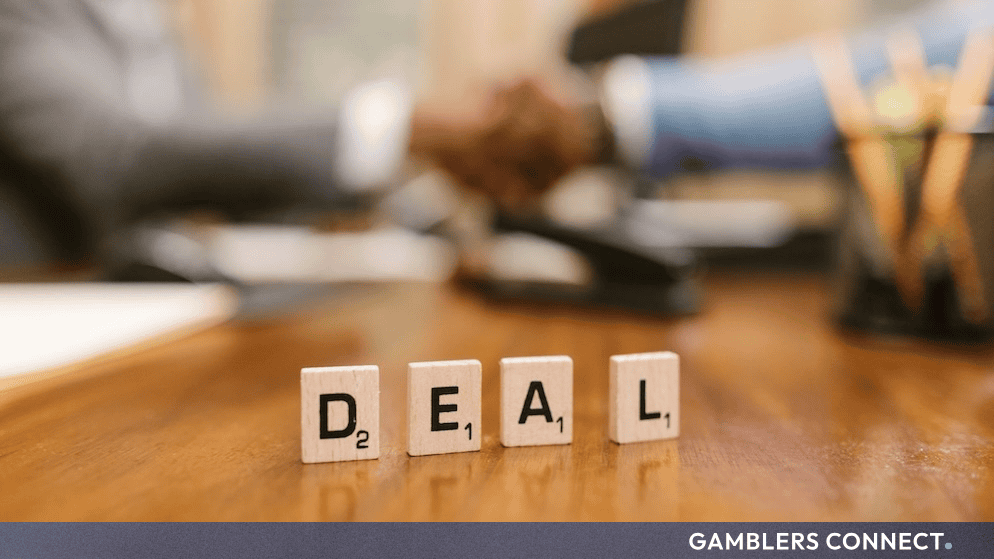 Wooden letter tiles spelling out the word "DEAL" on a boardroom table with a business handshake blurred in the background.