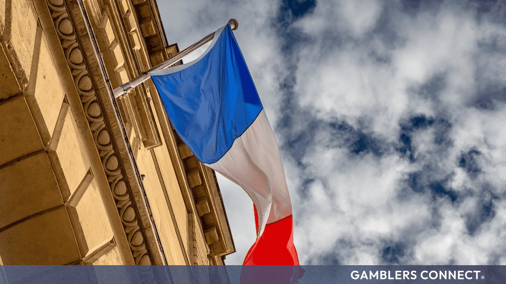 The French national flag waving proudly against a partly cloudy sky next to a classical stone building.