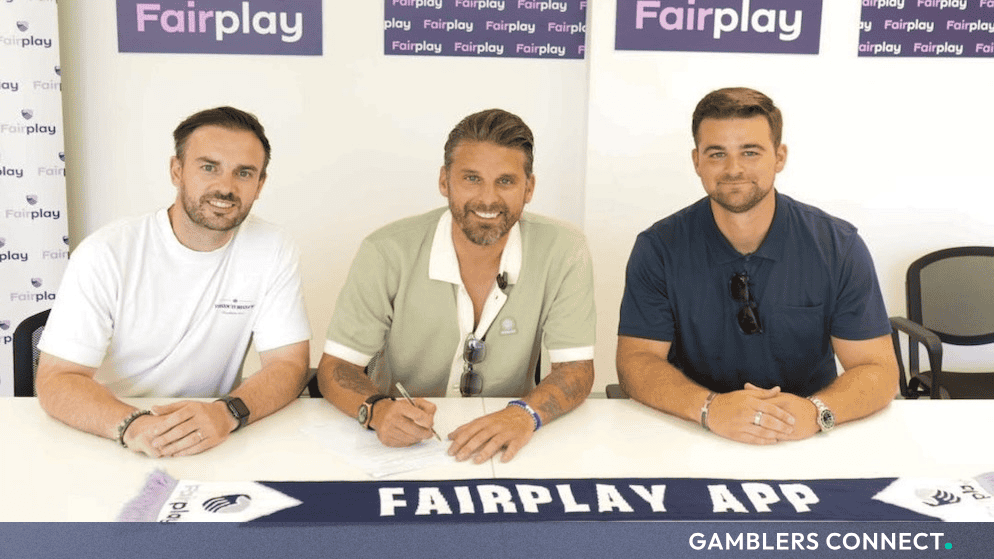 Former England footballer David Bentley seated at a desk flanked by executives, signing his ambassador contract with Fairplay alongside a branded club scarf.
